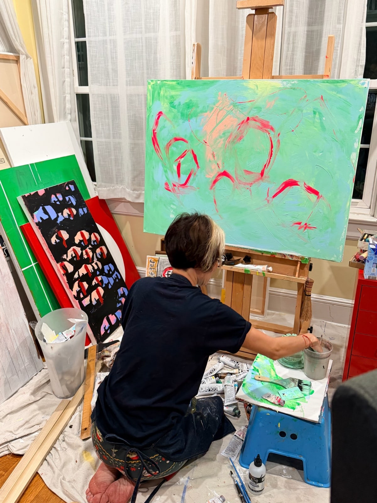 Chie Ushio kneeling on the floor of her studio at night, painting a large green and red canvas on an easel, surrounded by finished works and paint tubes.